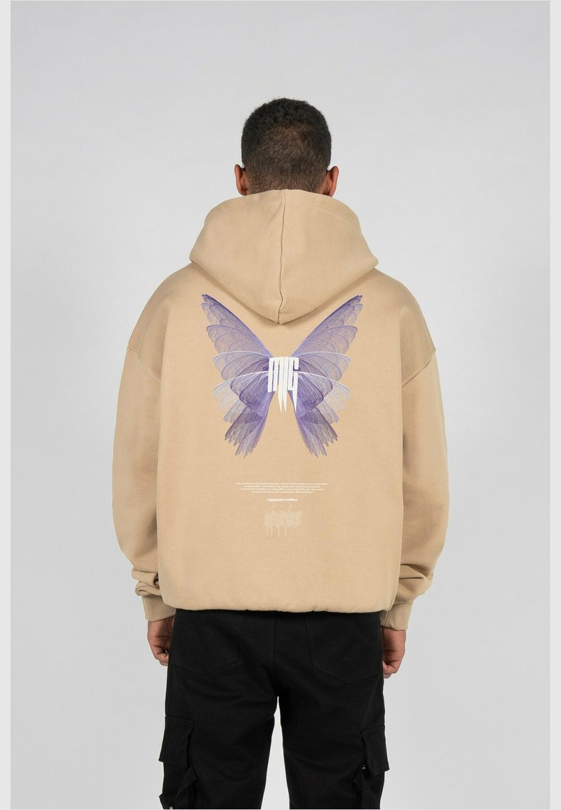 Homme portant un sweat à capuche beige avec un grand papillon violet imprimé dans le dos, associé à un pantalon cargo noir, debout devant un fond gris uni.