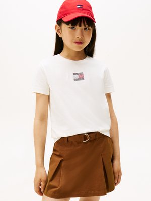 Witte T-shirt met een klein, kleurrijk logo op de borst, gecombineerd met een bruine rok met een riem en plooien; een rode pet maakt de outfit compleet.
