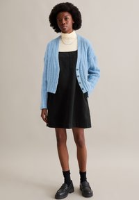 Hellblauer gerippter Cardigan über einem schwarzen Jeanskleid. Schwarze Loafer und Socken vervollständigen das Outfit. Einfaches Design mit minimalen Akzenten.