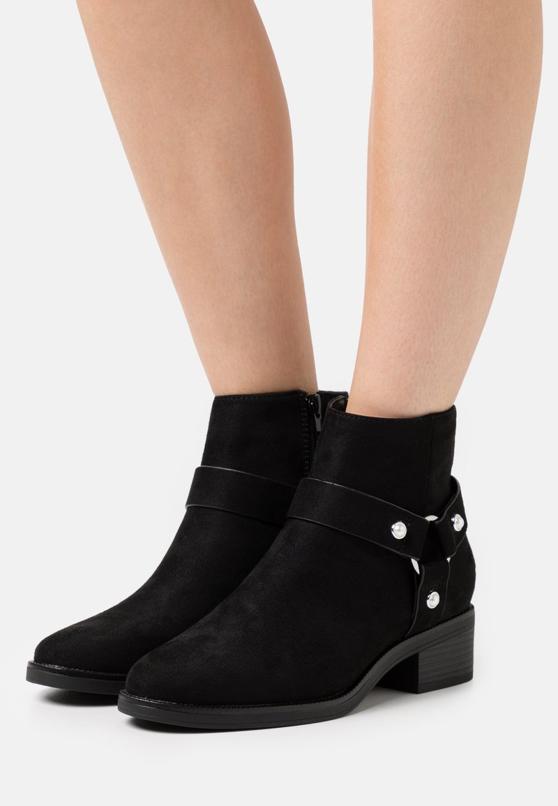 Botines de ante negro con punta redonda, tacón medio, cremallera lateral y correa decorativa con acentos plateados.