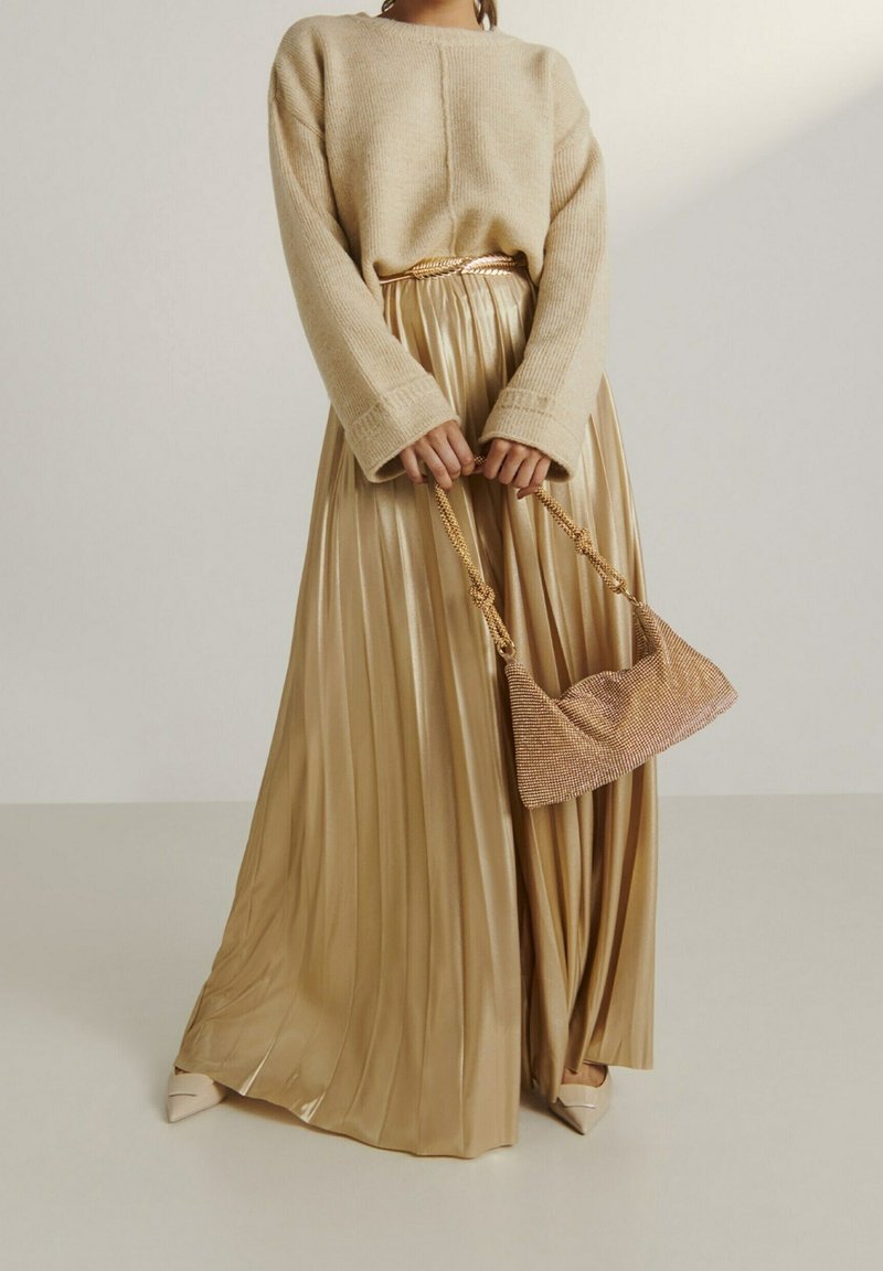 Beżowy sweter dziany z długimi rękawami, zestawiony ze złotą plisowaną maxi spódnicą. Dodatki obejmują małą metaliczną torbę oraz beżowe czółenka.