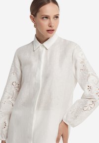 Wit linnen overhemd met een kraag, lange mouwen met ingewikkelde bloemrijke borduursels en een knoopsluiting aan de voorkant.