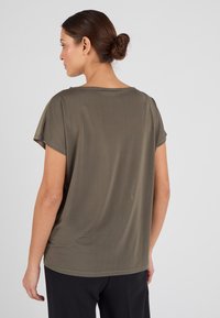 Mujer con cabello castaño recogido en un moño, vistiendo una blusa suelta de color verde oliva, vista desde atrás contra un fondo gris claro.