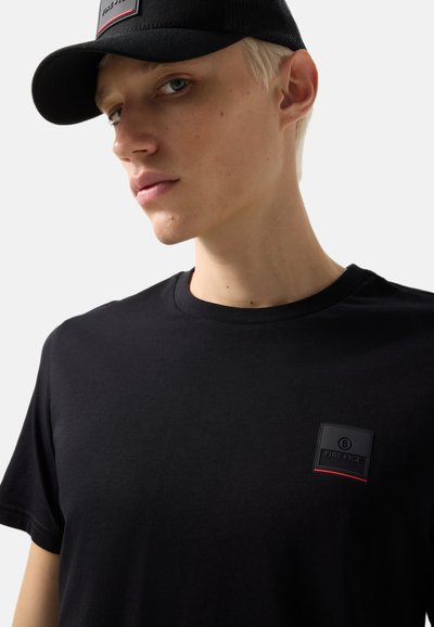 Ung person med kort blondt hår iført en sort kasket og en sort T-shirt med et lille "FIRE+ICE" logo-patch på brystet.