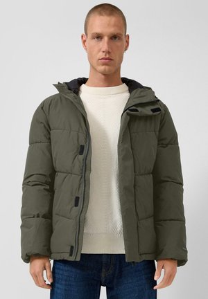 Uomo che indossa una giacca imbottita verde oliva sopra un maglione bianco lavorato a maglia e jeans blu su uno sfondo chiaro e uniforme.