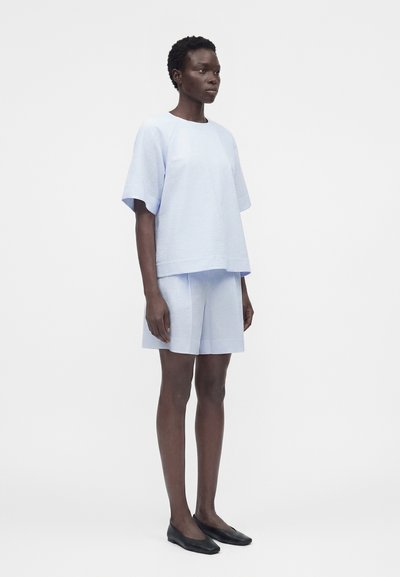 Γυναίκα που φοράει γαλάζιο υφασμάτινο κοντομάνικο τοπ και αντίστοιχα φαρδιά σορτς, με μαύρες μπαλαρίνες, σε απλό λευκό φόντο.