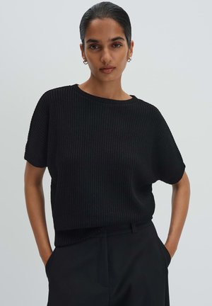 Donna con capelli scuri raccolti, che indossa una maglietta nera a maniche corte a coste e pantaloni neri, con le mani nelle tasche, su sfondo grigio neutro.