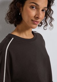 Dunkelbrauner Pullover mit Rundhalsausschnitt, der weiße Streifen an den Schultern hat. Weicher Stoff und lockere Passform.