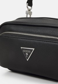 Borsa sintetica nera con zip argentata, logo triangolare "GUESS", texture liscia e design piatto. Accentuata da una chiusura metallica.