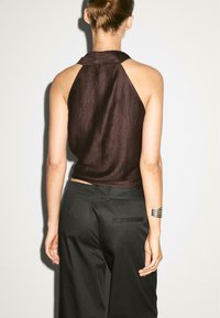 Modèle de dos, portant un haut sans manches marron foncé et un pantalon noir à jambes larges. Un bracelet en argent orne son poignet. Fond simple.