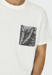 T-shirt bianca a maniche corte con taschino grigio e nero a motivo foglie di palma sul lato sinistro. Tessuto morbido e texturizzato.