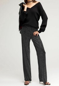 Zwarte geribbelde oversized trui met een V-hals, gecombineerd met glanzende zwarte wijde broek met een subtiele metallic glans. Zwarte hakken maken de look af.