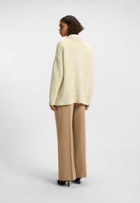 Maglione a maglia di colore crema, con texture e vestibilità ampia, maniche a costine e orlo asimmetrico, abbinato a pantaloni beige a gamba larga e tacchi.