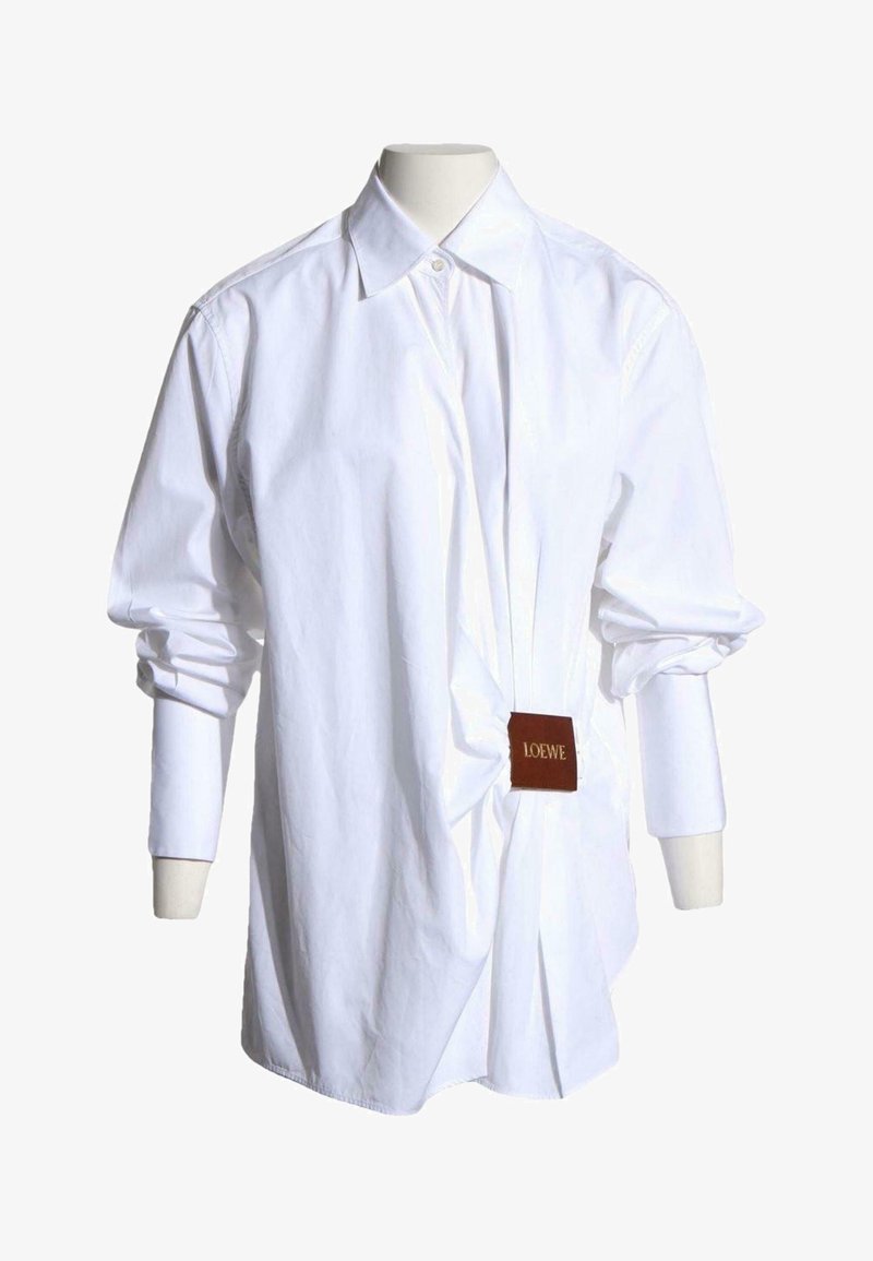 Chemise blanche à manches longues, boutonnée, avec un détail froncé à l’avant et une étiquette marron Loewe attachée sur un mannequin sans tête.