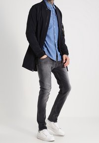 Zwarte bomberjack over een lichtblauw denim shirt, gecombineerd met grijze jeans en witte sneakers; casual, gelaagd outfit met verschillende texturen.
