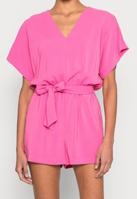 Vaaleanpunainen V-pääntie romper, jossa on lyhyet hihat, vyötäröllä vyö ja rento istuvuus. Valmistettu sileästä kankaasta, jossa on kevyt rakenne.