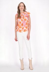 Blumenshirt mit Rüschensleeves in Orange- und Rosatönen, kombiniert mit weißen Skinny Jeans und beigen Stiefeletten. Glatte Stofftextur.
