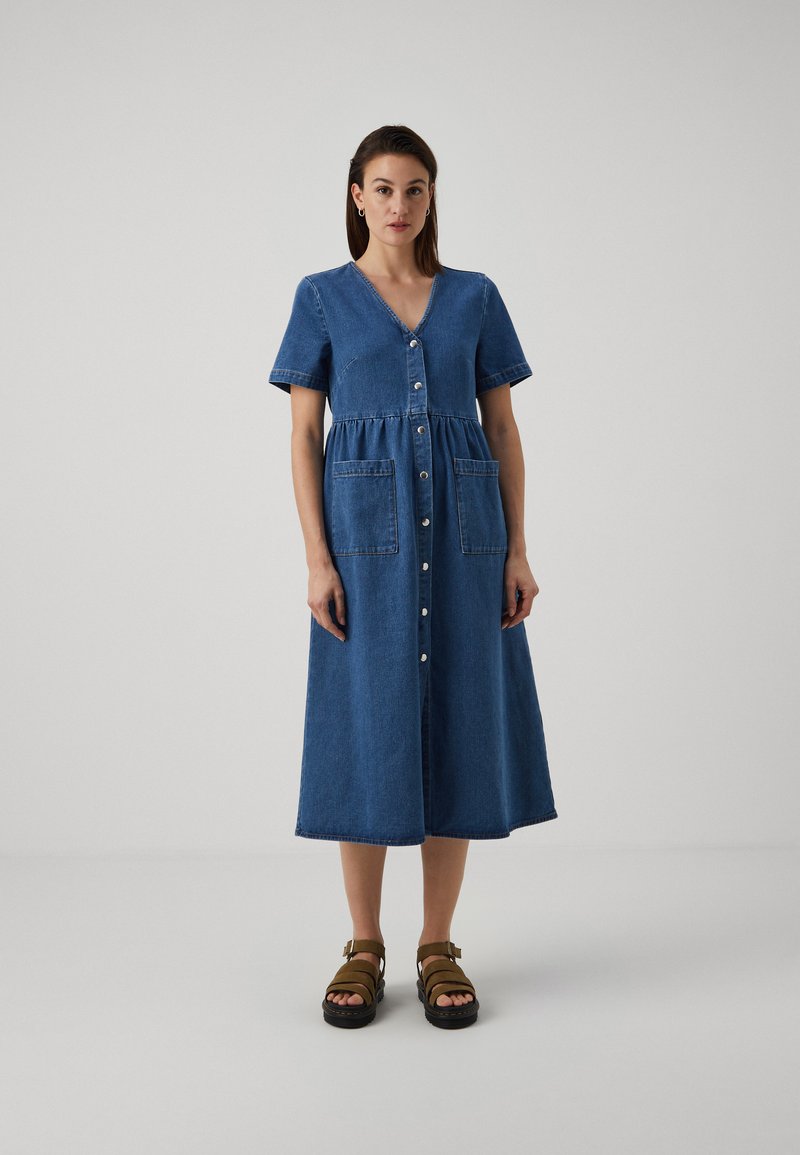 Denimkjole med korte ermer, V-hals, knappelukking foran og to påsydde lommer. A-linje silhuett når til midten av leggen. Mørk blå farge og myk tekstur.