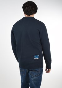 Navy blue sweatshirt with a round neckline, ribbed cuffs, and a stitched logo in bright blue at the lower back. Smooth texture.