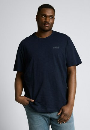 Mężczyzna w ciemnogranatowej koszulce z okrągłym kołnierzykiem i jasnoniebieskich dżinsach, stojący z jedną ręką w kieszeni na jednolitym tle.