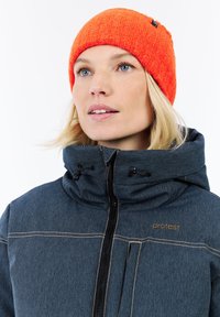 Giovane donna con occhi blu che indossa un cappellino di lana arancione brillante e una giacca blu con zip con il logo "protesta" sul petto.