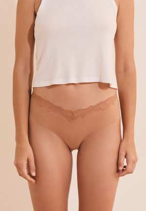 Femme portant des sous-vêtements couleur chair avec une bordure en dentelle et un débardeur court blanc, debout devant un fond beige avec les bras détendus le long du corps.