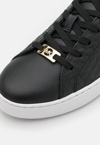 Schwarze Leder-Sneaker mit strukturierten Seiten, schwarzen Schnürsenkeln und einem goldenen Logoakzent am Ösenbereich. Weiße Gummisohle mit Stickdetails.
