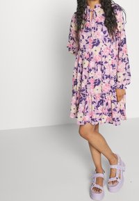 Šaty s květinovým vzorem v purpurové, růžové a krémové barvě. Mají dlouhé rukávy, volánkový lem a zavazovací výstřih. V kombinaci s fialkovými masivními sandály.