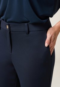 Pantalons bleu marine avec une texture lisse, dotés d'une poche avant, d'une fermeture à boutons et d'une ceinture. Le design ajusté met en valeur des lignes épurées.