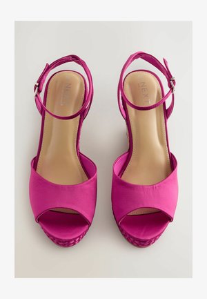 Svetlo roza sandale na visokem petnem delu z odprto konico in trakcem okoli gležnja, z vgrajenim tkanim platfromom, prikazane na nevtralni podlagi.