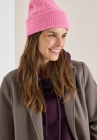 Gorro de punto rosa con textura acanalada, combinado con una sudadera morada oscura y un abrigo gris. El cabello es largo y ondulado. Fondo neutro.