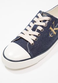 Marinblå kanvas sneakers med en vit gummitåspets, beiga snören och en guldfärgad logotyp på sidan. Har en texturerad sula för bättre grepp.