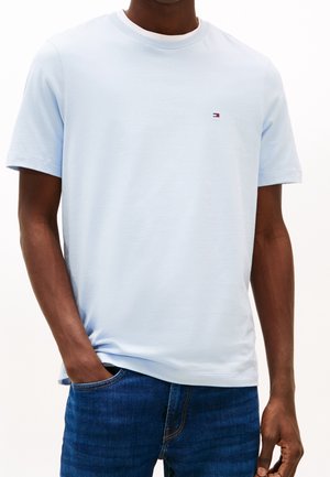 Mężczyzna w jasnoniebieskiej koszulce z okrągłym dekoltem z małym logo Tommy Hilfiger na piersi i niebieskich dżinsach, z jedną ręką w kieszeni.