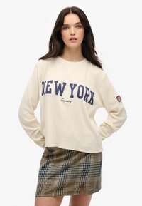 Felpa a maglia waffle di colore crema con la scritta "NEW YORK" in blu navy, abbinata a una gonna a quadri blu e marrone con spacco laterale.