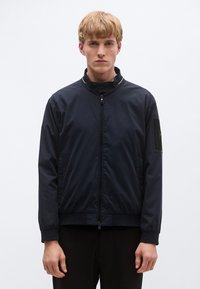 Tumesinine bomber-jakk kõrge kraega, ees tõmblukuga sulgemine, küljetaskud ja tõmblukuga relvatasku bränditud detailiga. Sile tekstuur.