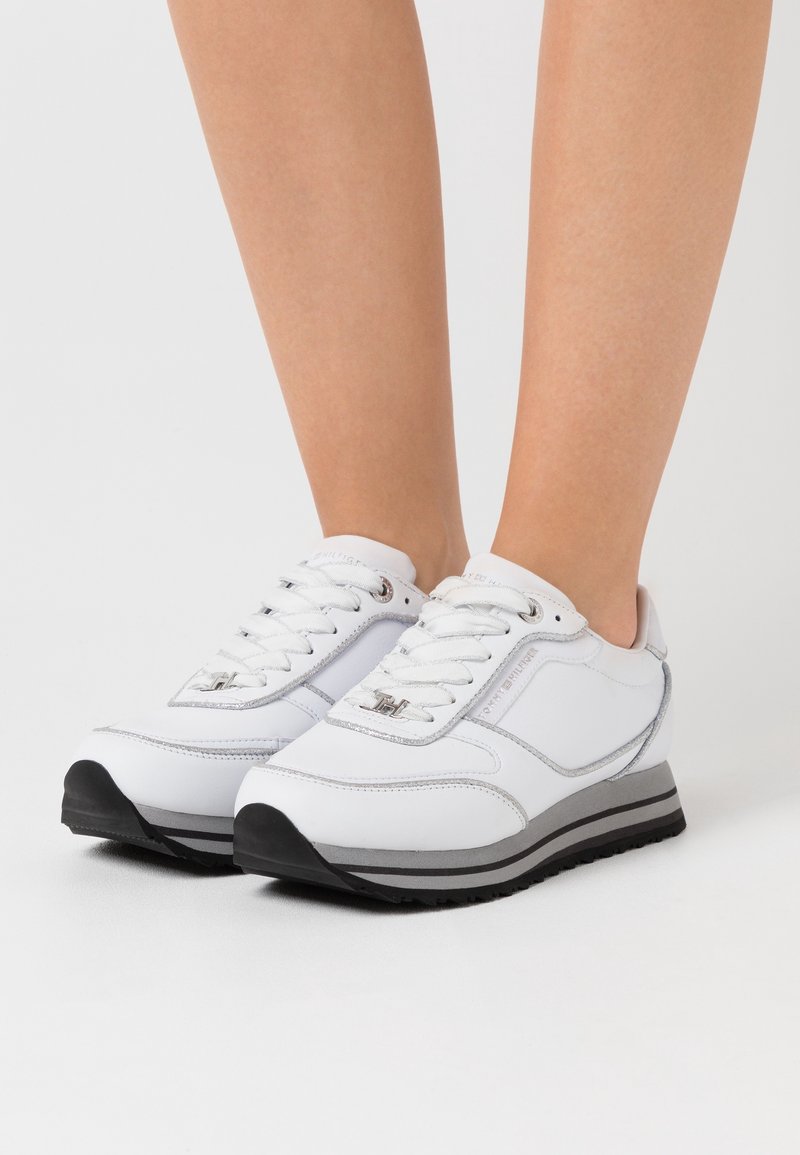 Baskets blanches présentant une tige en cuir lisse, un bout arrondi, des accents argentés et une semelle en caoutchouc noire avec un design stratifié.