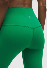 Zaļas legingi ar augstu jostasvietu, gludu tekstūru un pastiprinātiem šuvēm. Diskretā logo tuvu jostasvietai.