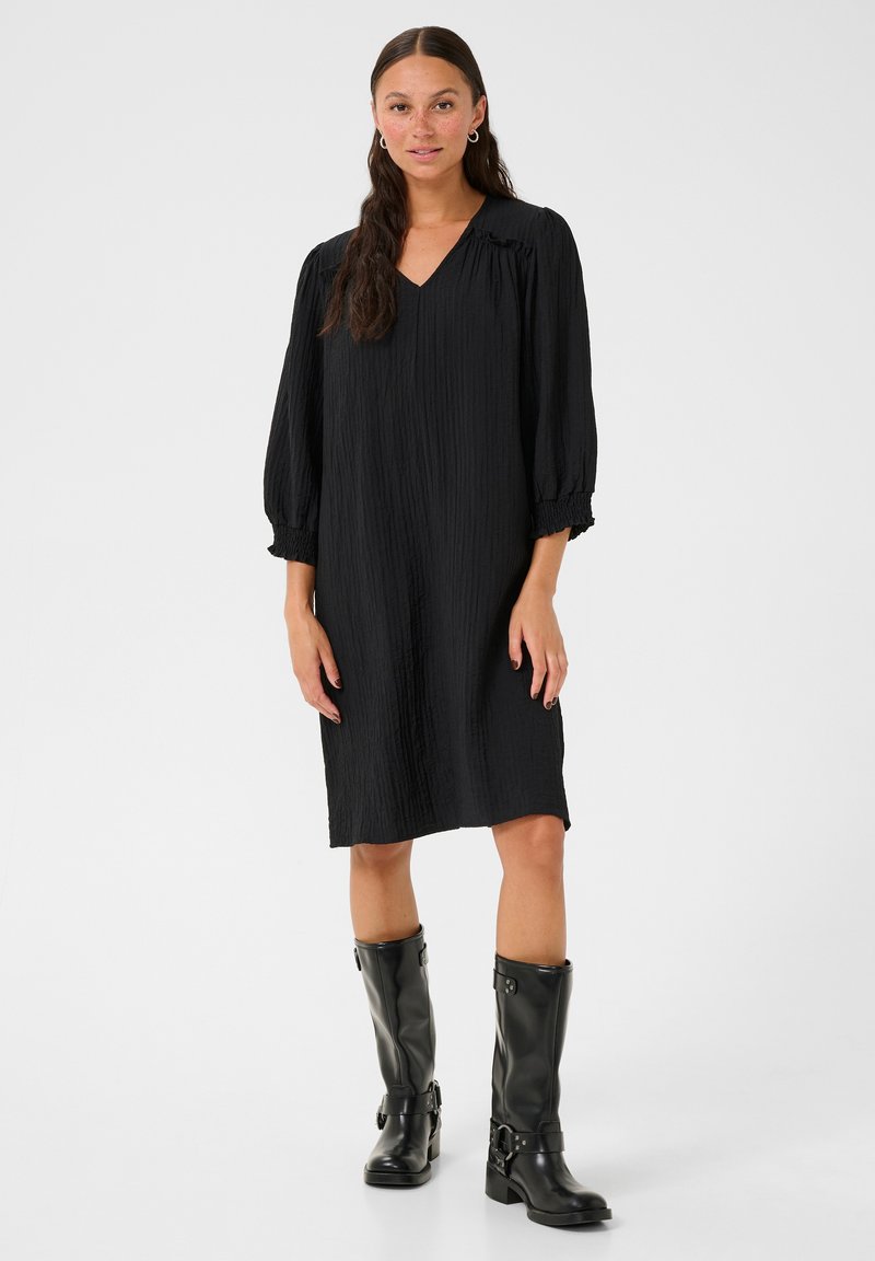 Robe noire, longueur genoux avec un décolleté en V et des manches bouffantes. Fabriquée en tissu texturé. Associée à des bottes noires brillantes hauteur genoux.