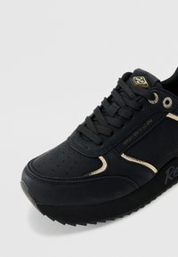 Zapatilla negra con un upper sintético suave, acentos dorados, detalles perforados, cordones redondos y una suela gruesa y texturizada.