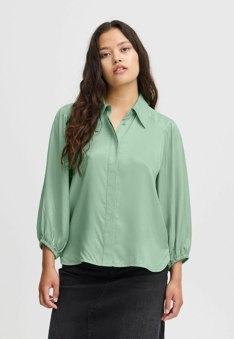 Blouse vert clair à manches longues avec un devant boutonné et des poignets élastiques. Tissu doux et lisse avec un ourlet arrondi et décontracté.