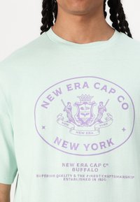 Mätovo zelené bavlnené tričko s fialovým potlačeným grafickým motívom, vrátane loga "New Era Cap Co" a textu "New York" s dekoratívnymi detailmi.