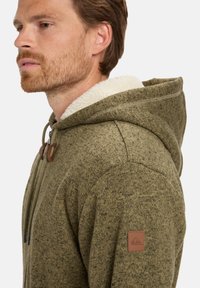 Groene hoodie met een gestructureerde gebreide stof, een zachte witte bontvoering in de capuchon en bruin leren accenten op de rits en linker schouder.