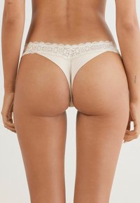 Beige string met kantafwerking, versierd met bloemmotieven, van een gladde stof, met een ondeugende snit, verlengd achterontwerp en verfijnde kantdetails op de taille.