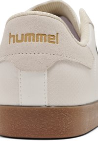 Adidași albi cu un spate din suede texturat, talpă din cauciuc și logo auriu. Prezintă o suprafață netedă și o parte din material cu un ușor model.
