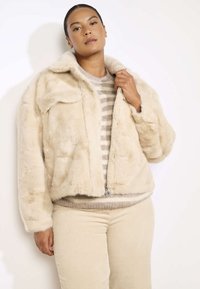 Bežna umetna krznena jakna z zadrgo spredaj, velikim ovratnikom in dvema sprednjima žepoma, oblečena čez svetlo prugasto pulover in bež kordne hlače.