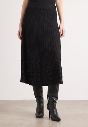 Jupe en maille côtelée noire avec un ourlet évasé, présentant des découpes circulaires en bas. Associée à des bottes noires à talons hauts.