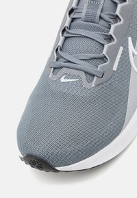 Chaussure de sport grise avec tige en mesh, surface texturée, logo blanc et semelle noire avec un motif de semelle.