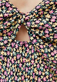 Zbliżenie czarnej tkaniny z małymi żółtymi, różowymi i białymi kwiatuszkami, zawiązanej w kokardę, z małym trójkątnym wycięciem ukazującym skórę pod spodem.