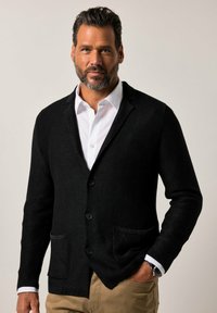 Schwarze Strickjacke mit zwei Fronttaschen, Fünf-Knopf-Verschluss und gerippten Bündchen. Kombiniert mit einem weißen Hemd mit Kragen und beigen Hosen.