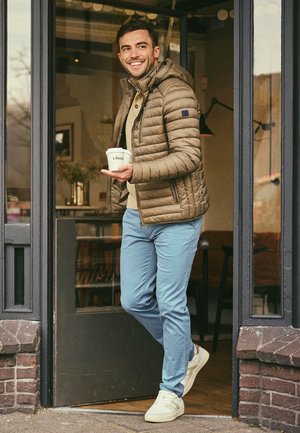 Pufferjack in lichtbruin, beige trui en blauwe broek. Witte sneakers. Houdt een koffiekop vast, staat bij een deur. Bakstenen entree.